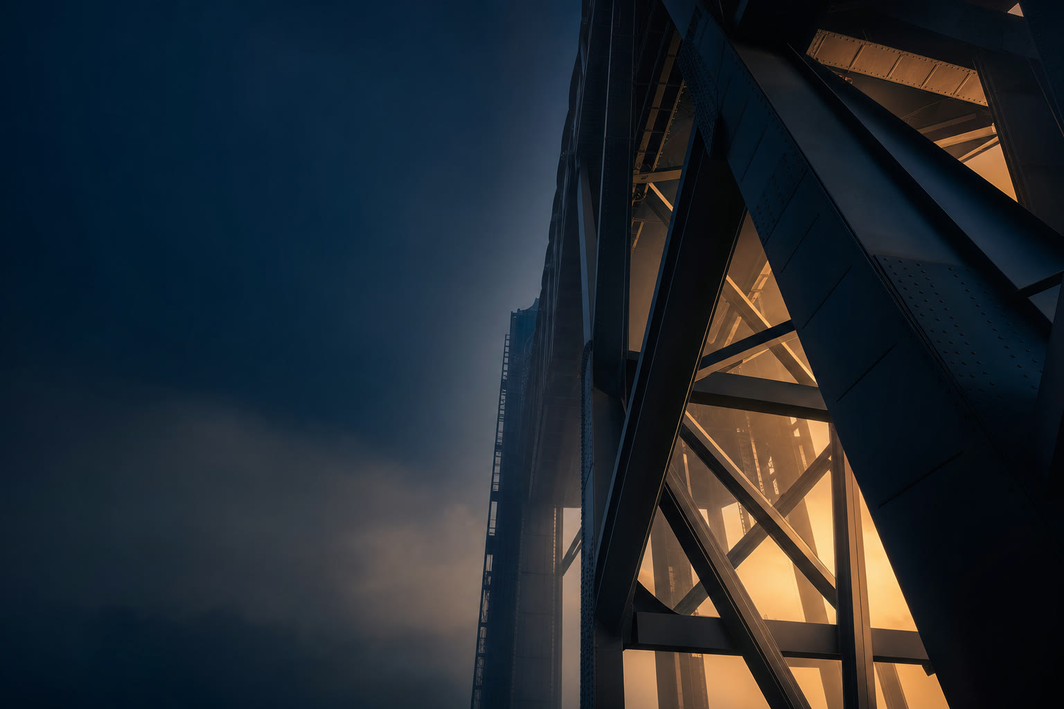 Steel girders and cross-bracing rising into a navy dusk sky, lit from within by warm golden light
